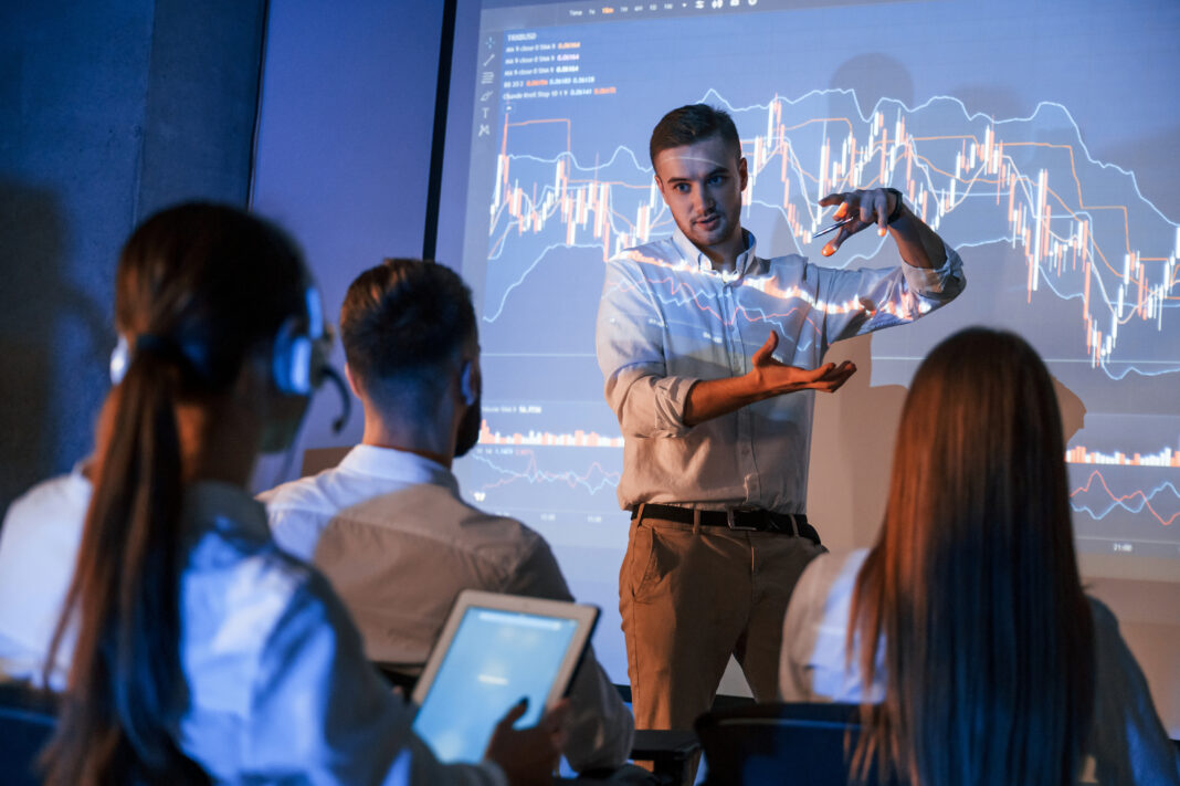 Company growth plan. Male leader talking to employees, showing information on the projector in office of stock exchange company. Copyright: standret - stock.adobe.com