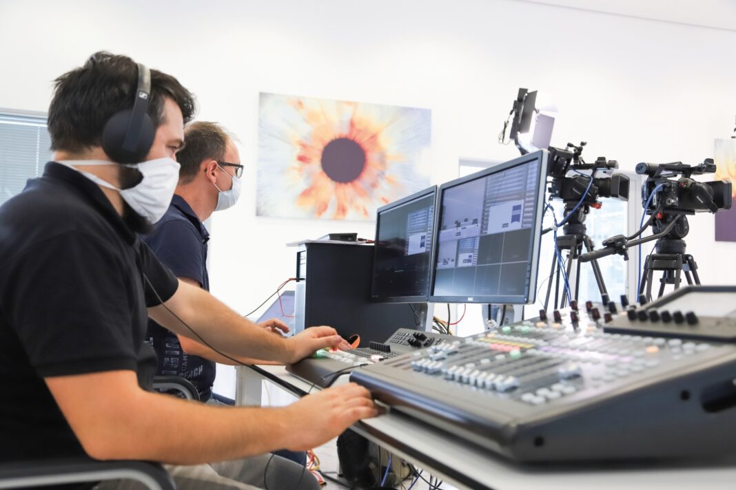 Zwei maskierte Männer arbeiten an Computermonitoren und Tonmischgeräten in einem Studio mit Kameras im Hintergrund.