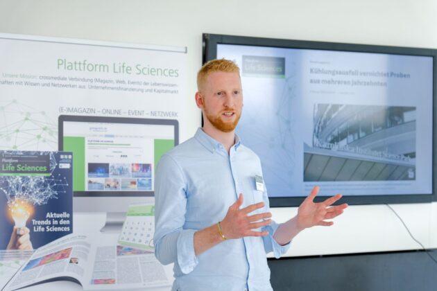 Das Bild zeigt eine Person mit roten Haaren, die eine Präsentation hält. Im Hintergrund sind zwei Displays zu sehen: Ein Banner mit dem Titel „Plattform Life Sciences“ und Text zu Magazin, Web, Events und Netzwerken im Bereich Life Sciences, sowie ein Bildschirm mit einem Artikel über einen Kühlungsfehler am „Karolinska Institutet“. Die Person steht neben den Displays und gestikuliert beim Sprechen.