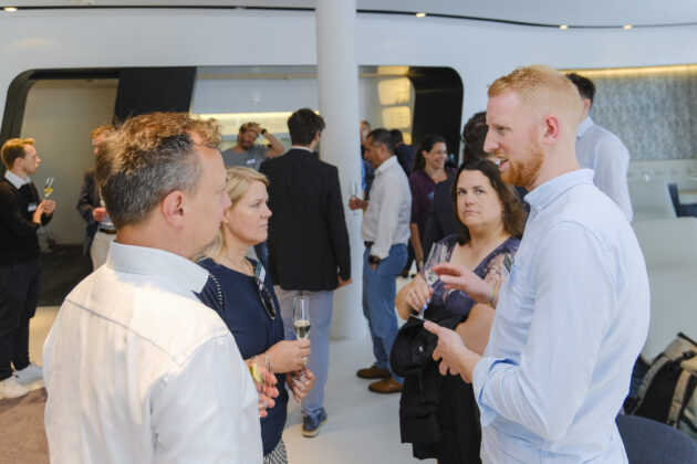 Menschen in Business-Casual-Kleidung unterhalten sich, einige mit Getränken. Moderne Umgebung mit stilvollem Design und Ambientebeleuchtung. Im Vordergrund: Mann in hellblauem Hemd, Frau im gemusterten Kleid, im Gespräch mit weiteren Personen. Andere Gäste im Hintergrund.