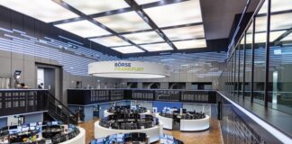Das Bild zeigt das Innere der Frankfurter Börse. Mehrere runde Handelstische mit Computermonitoren und Händlern sind zu sehen. Oben hängt ein großes Schild mit der Aufschrift „Börse Frankfurt“.