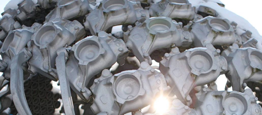 Das Bild zeigt eine Nahaufnahme einer Skulptur aus metallischen Autoteilen, die eine Kugelform bilden. Sonnenlicht erzeugt einen Lens-Flare-Effekt. Der Hintergrund ist unscharf, um die Details der Skulptur hervorzuheben.