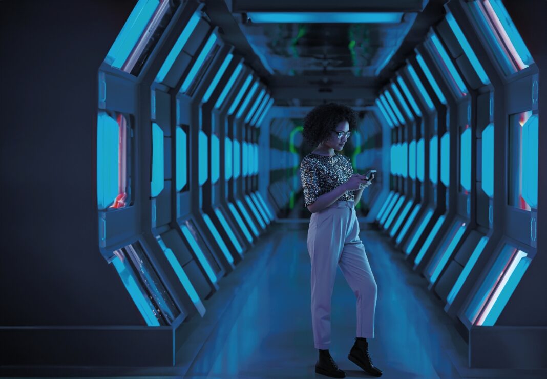 Das Bild zeigt eine junge Frau in einem futuristischen, blau beleuchteten Korridor. Sie trägt eine Brille, ein Paillettentop, hoch taillierte Hosen und schwarze Stiefeletten, während sie konzentriert auf ihr Handy schaut. Die Umgebung wirkt modern und technologisch fortschrittlich.