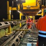 Arbeiter in orangefarbener Schutzkleidung und Helmen arbeiten nachts an einer Bahngleise. Schwere Maschinen und ein großes gelbes Gerüst sind sichtbar. Die Szene ist durch künstliche Lichter erhellt. Auf einer Jacke steht das Kürzel "FAS."
