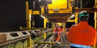 Arbeiter in orangefarbener Schutzkleidung und Helmen arbeiten nachts an einer Bahngleise. Schwere Maschinen und ein großes gelbes Gerüst sind sichtbar. Die Szene ist durch künstliche Lichter erhellt. Auf einer Jacke steht das Kürzel "FAS."