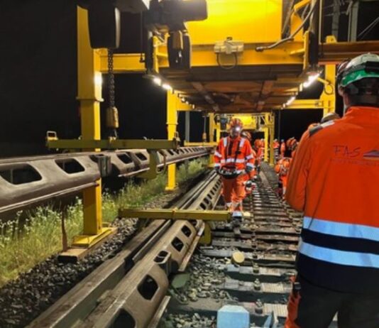 Vossloh bündelt mit Übernahme Kräfte in Frankreich Arbeiter in orangefarbener Schutzkleidung und Helmen arbeiten nachts an einer Bahngleise. Schwere Maschinen und ein großes gelbes Gerüst sind sichtbar. Die Szene ist durch künstliche Lichter erhellt. Auf einer Jacke steht das Kürzel "FAS."