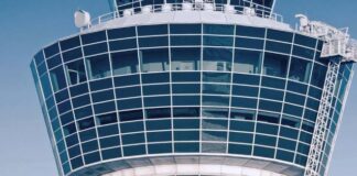 Das Bild zeigt den oberen Teil eines modernen Flughafen- Kontrollturms vor einem klaren blauen Himmel. Der Turm besteht überwiegend aus Glasfenstern, die in einem Gittermuster angeordnet sind und einen Panoramaausblick ermöglichen. Auf der Oberseite und an den Seiten sind verschiedene Antennen und technische Ausstattungen montiert.
