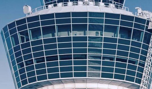 Frequentis: Übernahme der Groiss Informatics GmbH Das Bild zeigt den oberen Teil eines modernen Flughafen- Kontrollturms vor einem klaren blauen Himmel. Der Turm besteht überwiegend aus Glasfenstern, die in einem Gittermuster angeordnet sind und einen Panoramaausblick ermöglichen. Auf der Oberseite und an den Seiten sind verschiedene Antennen und technische Ausstattungen montiert.