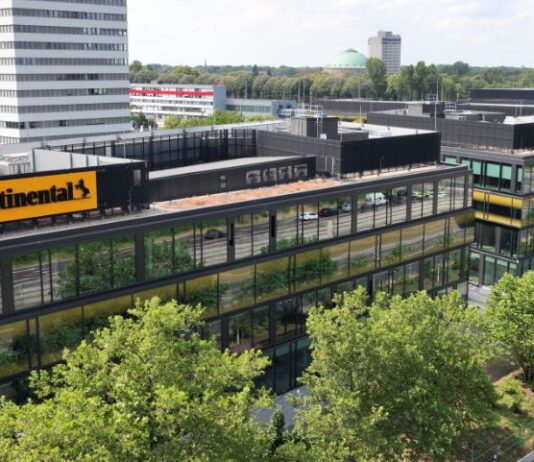 Continental prüft Ausgründung der Automotive-Sparte Das Bild zeigt ein modernes Bürogebäude mit dem „Continental“-Logo. Umgeben von Glasgebäuden, Grünflächen und Bäumen, ist im Hintergrund ein hoher weißer Bau und ein entferntes Wahrzeichen mit grüner Kuppel sichtbar. Eine Straße verläuft entlang des Komplexes, mit einigen Fahrzeugen, und der Himmel ist teils bewölkt.
