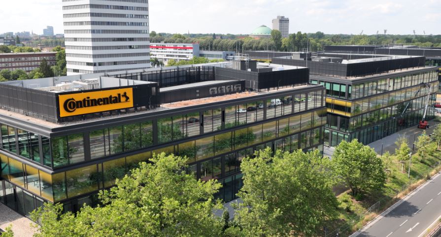 Das Bild zeigt ein modernes Bürogebäude mit dem „Continental“-Logo. Umgeben von Glasgebäuden, Grünflächen und Bäumen, ist im Hintergrund ein hoher weißer Bau und ein entferntes Wahrzeichen mit grüner Kuppel sichtbar. Eine Straße verläuft entlang des Komplexes, mit einigen Fahrzeugen, und der Himmel ist teils bewölkt.
