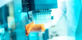 Das Bild zeigt ein Labor mit einem automatisierten Gerät zur Handhabung von Proben. Eine Hand in einem orangefarbenen Handschuh interagiert mit einem rechteckigen Behälter. Der Hintergrund ist mit Laborgeräten und -materialien in bläulichem Licht gefüllt. Copyright: Medigene AG
