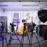 Das Bild zeigt ein professionelles Video-Set mit Kameras auf Stativen, einer gelben Sessel, Studioleuchten und Postern von „Straight Outta Compton“ und „The Night Manager“. Weitere Ausrüstung wie Kabel und Elektronik ist ebenfalls sichtbar.