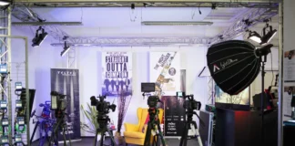 Das Bild zeigt ein professionelles Video-Set mit Kameras auf Stativen, einer gelben Sessel, Studioleuchten und Postern von „Straight Outta Compton“ und „The Night Manager“. Weitere Ausrüstung wie Kabel und Elektronik ist ebenfalls sichtbar.