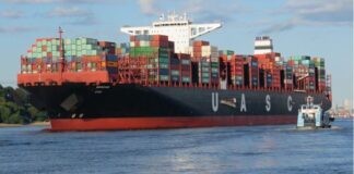 Das Bild zeigt ein großes Containerschiff mit den Buchstaben „U A S C“ und bunten Containern auf dem Deck. Ein kleineres Schiff fährt davor, während sich das Ganze auf einem Wasserweg mit grünem Ufer und Gebäuden im Hintergrund abspielt.