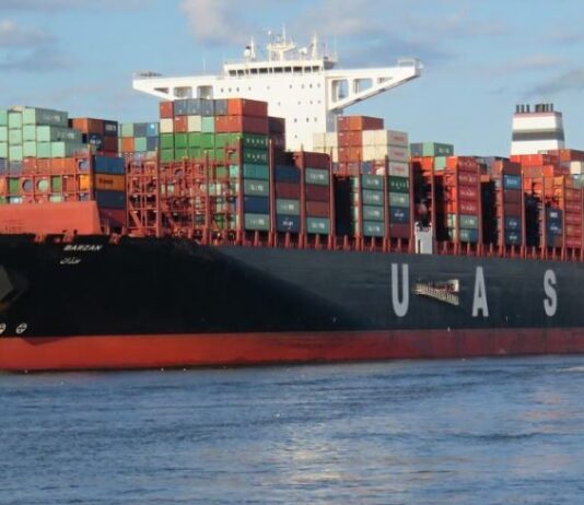 INDUS: Neue Beteiligung im Engineering-Segment Das Bild zeigt ein großes Containerschiff mit den Buchstaben „U A S C“ und bunten Containern auf dem Deck. Ein kleineres Schiff fährt davor, während sich das Ganze auf einem Wasserweg mit grünem Ufer und Gebäuden im Hintergrund abspielt.