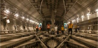 Das Bild zeigt eine Szene in einem großen unterirdischen Tunnel. Mehrere Bauarbeiter, die orange und gelbe reflektierende Sicherheitswesten und Helme tragen, sind im Tunnel verteilt und mit verschiedenen Aufgaben beschäftigt. Der Tunnel wird von zahlreichen Lichtern entlang der Wände beleuchtet, und in der Mitte verlaufen mehrere Bahngleise. An den Seiten sind Kabel und andere Baumaterialien zu sehen, die zu einem aktiv bearbeiteten Infrastrukturprojekt gehören.