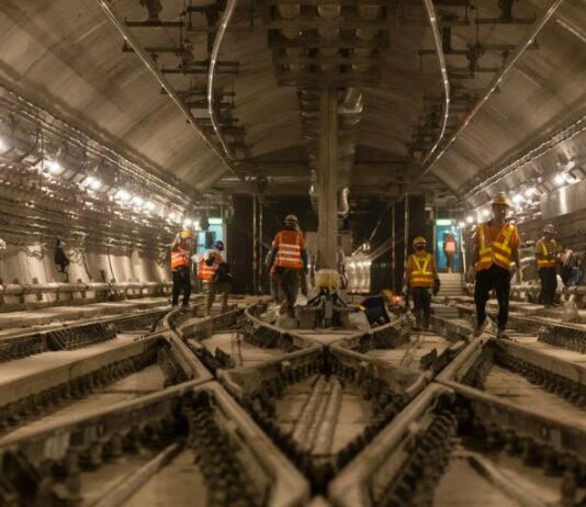Knorr-Bremse: Kauf von Alstom Signaling Nordamerika Das Bild zeigt eine Szene in einem großen unterirdischen Tunnel. Mehrere Bauarbeiter, die orange und gelbe reflektierende Sicherheitswesten und Helme tragen, sind im Tunnel verteilt und mit verschiedenen Aufgaben beschäftigt. Der Tunnel wird von zahlreichen Lichtern entlang der Wände beleuchtet, und in der Mitte verlaufen mehrere Bahngleise. An den Seiten sind Kabel und andere Baumaterialien zu sehen, die zu einem aktiv bearbeiteten Infrastrukturprojekt gehören.