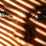 Close-up of a woman's face partially obscured by window blinds casting stripes of light and shadow. Copyright: musa - stock.adobe.com