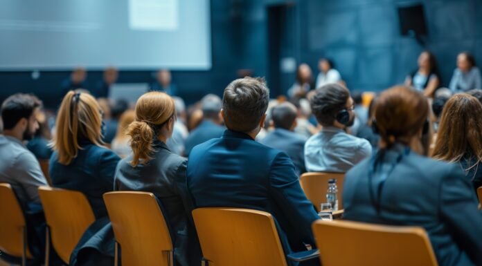 Menschen sitzen auf Stühlen, man sieht ihren Rücken. Sie schauen auf eine Leinwand.