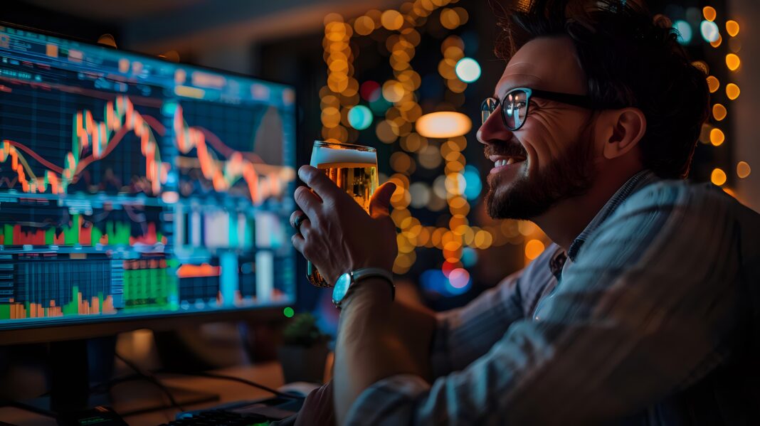 Successful male trader in glasses celebrating with beer while monitoring stock market charts on multiple screens, perfect for finance, investment and trading success concepts.