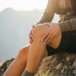 Case Study: BIONCaRT – Stammzellen für die Regeneration geschädigter Gelenke Symbolbild. Closeup of a hiker holding their knee in pain while sitting on a rock during a mountain hike at sunset. Copyright: ViskBx - stock.adobe.com