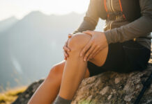Symbolbild. Closeup of a hiker holding their knee in pain while sitting on a rock during a mountain hike at sunset. Copyright: ViskBx - stock.adobe.com