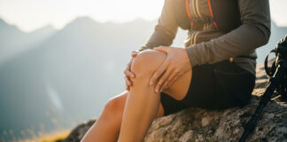 Symbolbild. Closeup of a hiker holding their knee in pain while sitting on a rock during a mountain hike at sunset. Copyright: ViskBx - stock.adobe.com